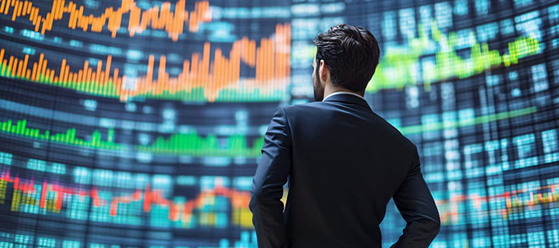 A man in a suit seen from behind, looking at a wall of stock charts and price tables
