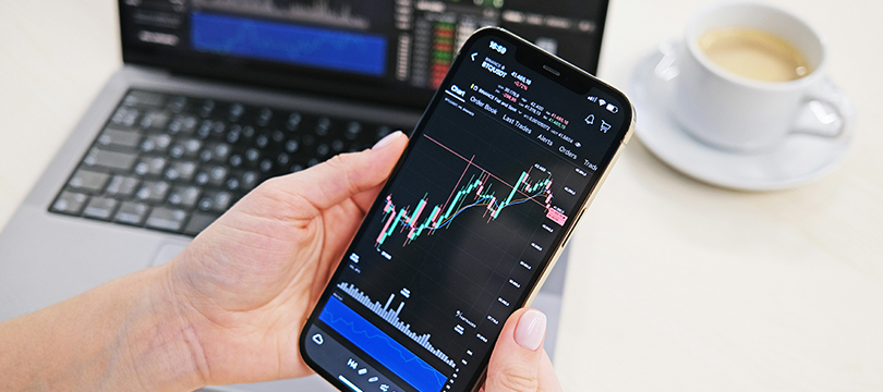 Hands holding a smartphone showing a candlestick chart in front of a laptop and a cup of coffee
