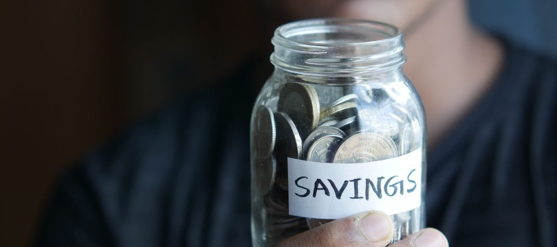 A hand holding up a glass jar partly filled with coins, with a handwritten label reading SAVINGS, in front of a person in a dark T-shirt