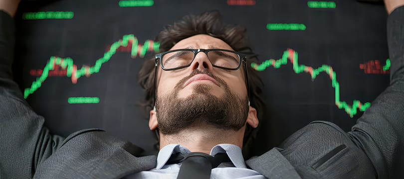 A man in a shirt and tie lying back with his eyes closed and hands behind his head in front of a screen of price charts